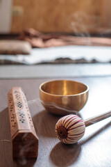 Decorative wooden incense holder releasing smoke next to a golden singing bowl and a striped mallet on a wooden surface, creating a serene atmosphere for meditation and relaxation
