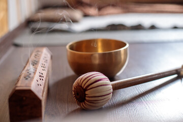Wooden incense holder emitting soft smoke beside a golden singing bowl and a striped mallet on a...