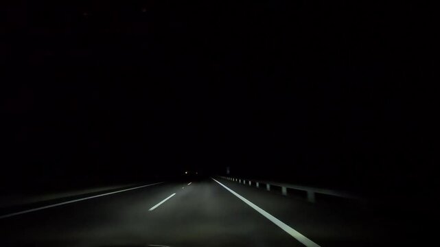 Nighttime highway drive, view from a car. Dark road illuminated by headlights, leading to a well-lit section and an exit ramp toward the end. Smooth, atmospheric journey.