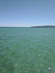 Vividly sparkling blue and green island water under a bright, clear sky, with sunlight glistening off the surface like scattered diamonds. The hues of the water range from deep azure to emerald.