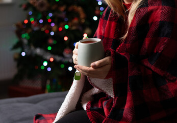 Close up of a woman holding a mug with hot drink and sitting on a sofa with a red blanket