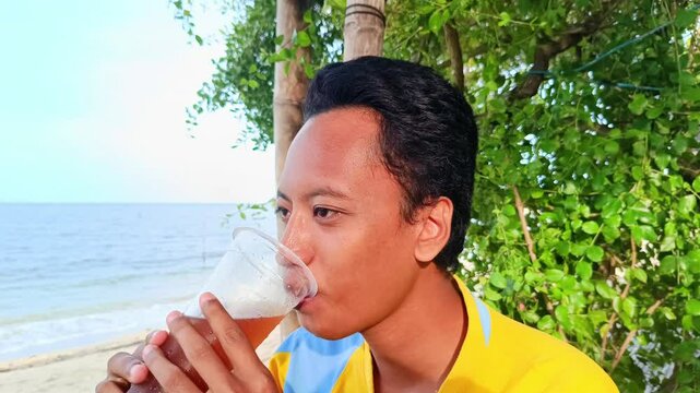 A asian guy is enjoying drinking iced tea with a beach background when summer vacation. Slow motion bit . Focus on man, 