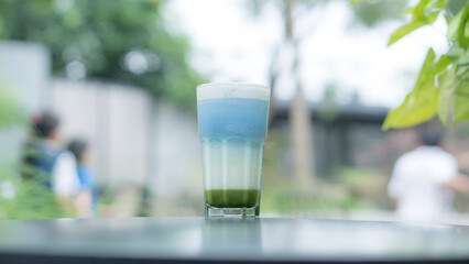 A glass of colorful two-tone drink, possibly a cocktail or refreshing beverage, sits on a textured table, outdoors. The drink has two distinct layers of color, a purple top and an orange bottom