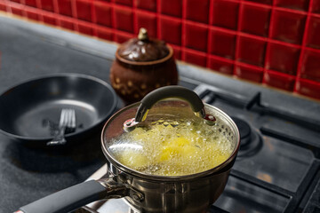 Potatoes cooked in a saucepan in water with a bay leaf under a lid. View of a stainless steel gas cooktop on a black table with red ceramic tiles on the wall.
