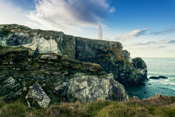 Fototapeta premium A lighthouse stands tall on a rocky cliff overlooking the ocean, surrounded by grassy terrain. The sky is partly cloudy, adding a serene atmosphere to the coastal setting.