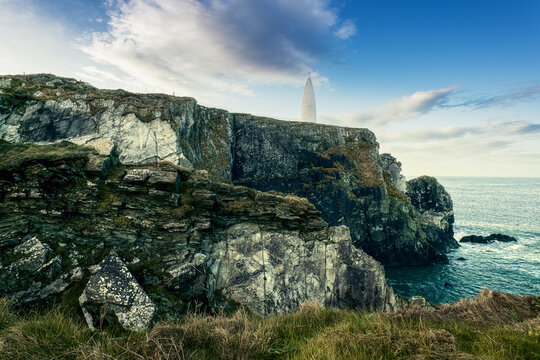 A lighthouse stands tall on a rocky cliff overlooking the ocean, surrounded by grassy terrain. The sky is partly cloudy, adding a serene atmosphere to the coastal setting.