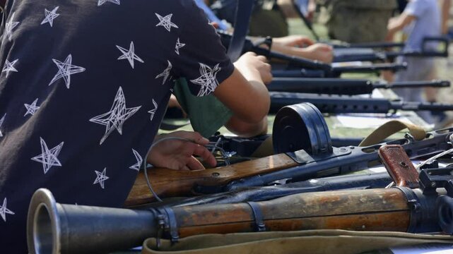 A group of curious children gathered around various military weapons, including a submachine gun, machine gun, and a handheld anti-tank grenade launcher, showcasing their interest in technology.