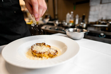 A chef sprinkles grated cheese over a gourmet dish in a professional kitchen. The focus is on the art of plating and the attention to detail in culinary presentation.
