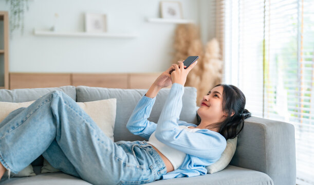 Happy cheerful positive young Asian woman on the phone at home, smiling girl using app and browsing on the internet on smartphone lying on sofa