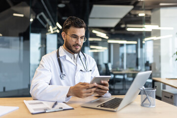 Mature man doctor seated at a desk, interacting with a tablet and laptop to access medical records,...
