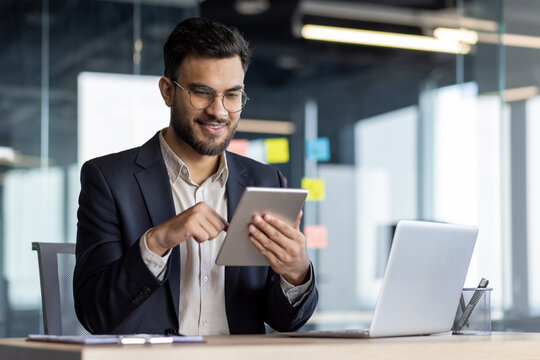 A mature man in formal business attire works confidently using a tablet device. He is seated at his desk in an office with a relaxed yet focused demeanor, surrounded by work tools.
