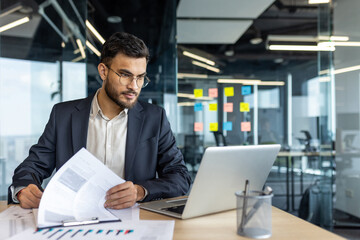 Mature man businessman sits at a desk, analyzing reports and working on a laptop. The professionally dressed individual is based in a bright, modern office with an organized workspace.