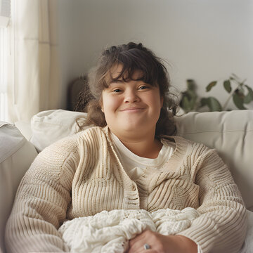 A warm and natural portrait of a young woman with Down syndrome sitting comfortably on a white sofa, wearing a cozy beige cardigan, and smiling softly in a bright, serene living room.
