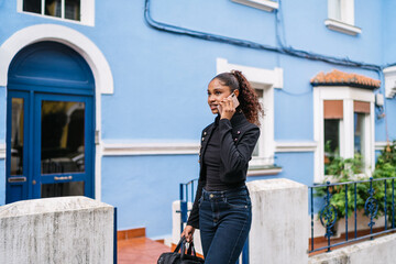 Young latin woman walking and talking on smartphone in front of blue building