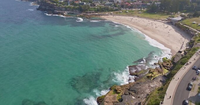 Zoom-in aerial view of Bronte Beach in Sydney, Australia, showing turquoise ocean water, golden sand, rolling waves, and scenic Pacific coastal landscapes in a smooth cinematic motion.