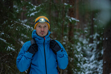 Male hiker, equipped with a headlamp and gloves, navigating a snowy forest trail during winter,...