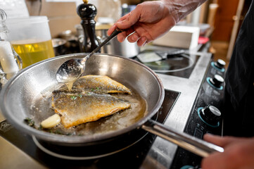 A chef is frying fish in a stainless steel pan, using a spoon to baste it with oil and herbs. The...