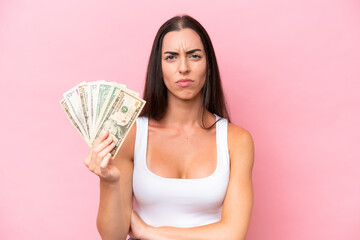 Young caucasian woman taking a lot of money isolated on pink background with sad expression