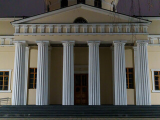 Fototapeta premium The strong and tall classical columns stand proud in the nighttime. The warm light accentuates the entrance of the building, creating a calm atmosphere in the surroundings.