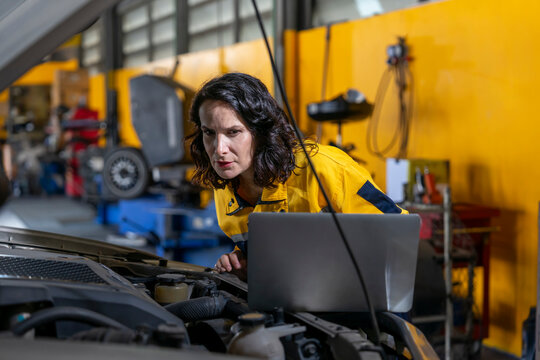 woman middle aged experienced mechanic in uniform using laptop examining car engine,concept of garage service,business,industry
