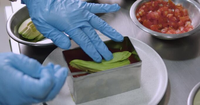 hands wearing blue gloves putting a layer of avocado slices during the preparation of Peruvian Causa
