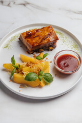 Glazed pork belly, crispy roasted potato wedges, fresh basil leaves, grated cheese, and vibrant tomato sauce elegantly arranged on a white plate. Restaurant or cafe menu, cookbook recipe
