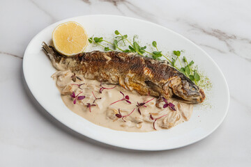 Delicious grilled fish served on a white plate with creamy sauce, lemon slice, microgreens, and red sprouts, creating an appetizing culinary presentation. Restaurant or cafe menu, cookbook recipe