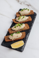 Creamy forshmak spread atop toasted rye bread slices, garnished with fresh chives and a lemon wedge, creates an elegant culinary scene on a black stone board. Restaurant or cafe menu, cookbook recipe