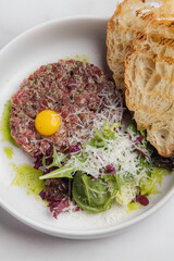 Classic steak tartare is presented with a bright egg yolk, parmesan cheese, fresh salad, and toasted bread slices on a white plate, set on elegant marble. Restaurant or cafe menu, cookbook recipe
