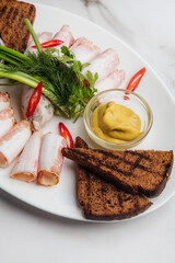 Thinly sliced ukrainian salo served with mustard, horseradish, rye bread, green onions, dill and red chili peppers arranged on white oval plate. Restaurant or cafe menu, cookbook recipe