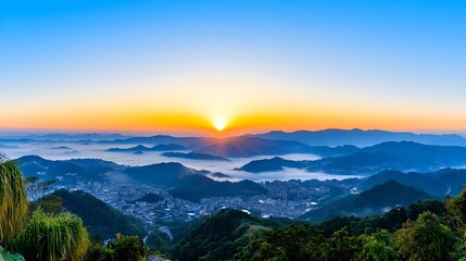 Fototapeta premium Majestic Sunrise Over Mountain Range and Valley, City Below Covered in Mist, Radiant Sky with Golden Orange Glow, Blue Horizon, and Green Hills in a Serene Scenic Landscape View