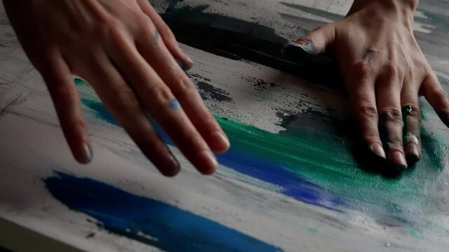 Close-up shot showcases an artist applying vibrant green and blue oil paint onto a canvas with her hands. Rings embellish her fingers, adding elegance to the artistic expression
