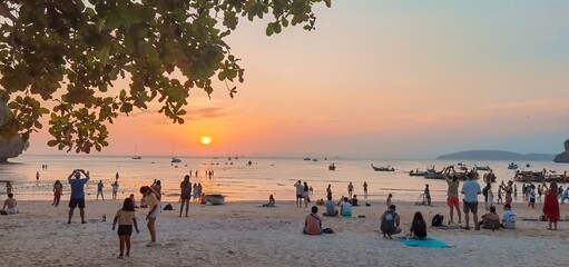 Panoramic photo of a Thai beach with people watching the warm and colorful sunset.