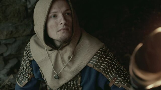 Medieval warrior dressed in chainmail armor with hood, holding lit torch and gazing up as exploring fortress tunnel. View from above