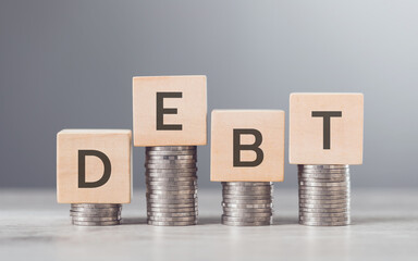 Stack of coins neatly arranged on top of a wooden block labeled "DEBT," symbolizing financial burdens and the weight of monetary obligations, set against a neutral background to emphasize the concept
