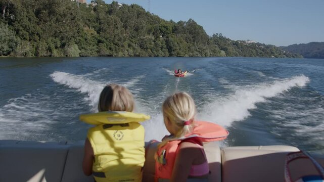 Two children in life jackets watch family tubing from a motorboat on a sunny lake, enjoying a fun summer day