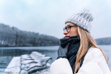 Woman wearing glasses and cozy winter attire, smiling, enjoying a snowy landscape by the lake
