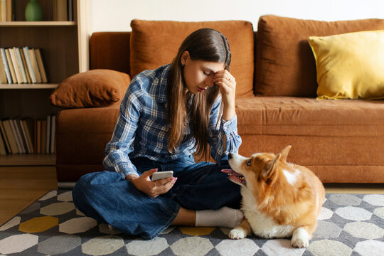Upset woman using smartphone sitting beside Welsh Corgi dog on floor carpet in living room, pet looking at lady