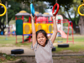 Happy girl hanging on monkey bar by hand doing exercise. Little Asian girl playing at outdoor playground in the park on summer vacation. Healthy activity.