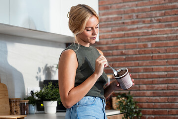 Beautiful young woman eating chocolate ice cream in the kitchen at home
