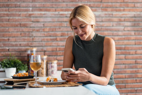 Pretty young woman eating sushi while using smartphone in the kitchen at home