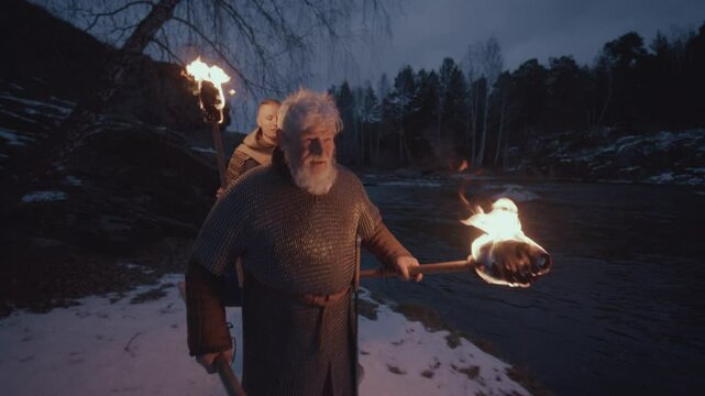 Two armored warriors in medieval chainmail and cloaks, holding burning torches and axe as walking together along snow-covered riverbank at night