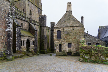 Une petite place de Locronan, les pavés mouillés reflètent la lumière sous un ciel gris. À droite, les murs de l'église se dressent, tandis qu'en face, une maison en pierre complète l'atmosphère paisi