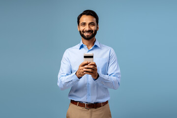 Fototapeta premium Portrait of smiling, happy bearded middle aged Indian man holding mobile phone, using mobile app