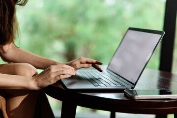 Young woman working on a laptop, focused and engaged, in a cozy indoor setting with natural light and lush green background, representing productivity and relaxation