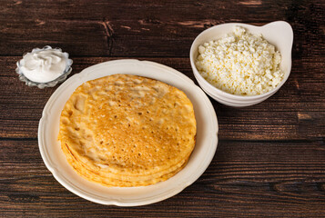 Pancakes are on a white plate, next to it is a bowl of sour cream and cottage cheese on a wooden table