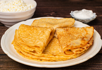 Pancakes are on a white plate, next to it is a bowl with sour cream and cottage cheese on a wooden table