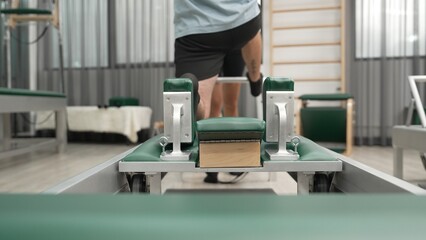 Man practicing kneeling stretch on pilates reformer with assistance to improve flexibility and strength. Trainer assist student doing lunge stretch on the reformer for enhancing balance. Habituate.