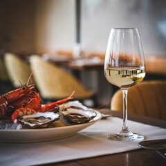 A photo of a close-up shot of a table in a cozy restaurant. There is a glass of white wine in the foreground, next to a plate of appetizing seafood