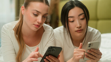 Two happy smiling multiracial friends multiethnic women diversity girls female Caucasian Asian Japanese Chinese relaxing on bed together using mobile phones discuss news online social media talking